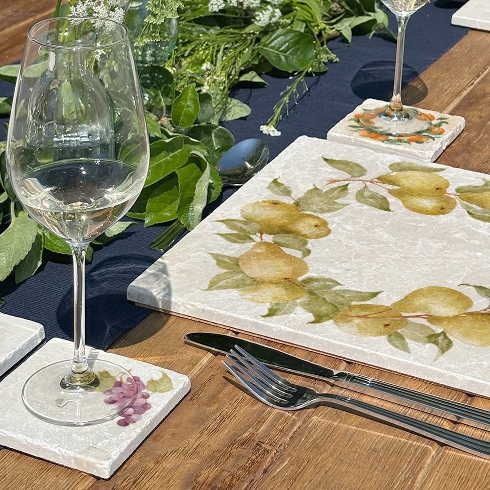 A farmhouse dining table set for a dinner party with marble placemats and coasters. The coasters and placemats have mix and match designs featuring different fruits.