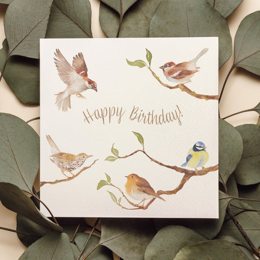 A greetings card laid flat on a table on top of dark green leaves. The greetings card reads Happy Birthday in brown text surrounded by garden birds on branches in a watercolour style.