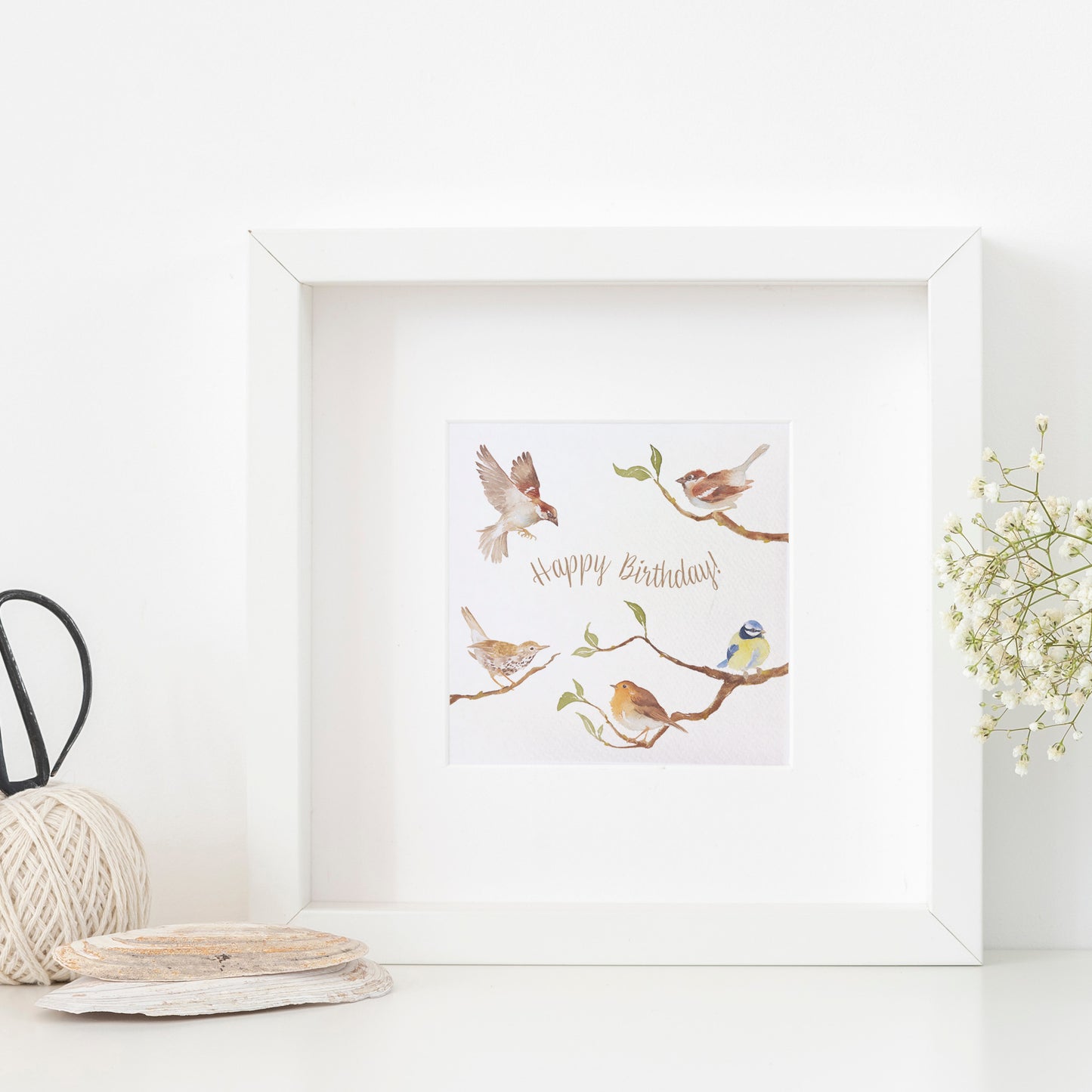 A greetings card displayed as an art print in a white frame propped up on a shelf next to a plant. The card reads Happy Birthday in brown text surrounded by garden birds on branches in a watercolour style.