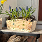 An arched wooden planter box full of spring flowers. The planter shows a watercolour repeat garden pattern design including welly boots, vegetables, gardening gloves, garden spade, snails, bees, robins and secateurs. A wonderful gift for an avid gardener.