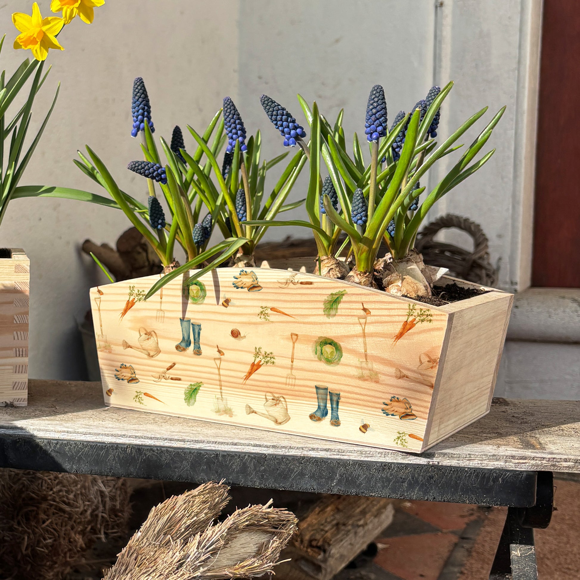 An arched wooden planter box full of spring flowers. The planter shows a watercolour repeat garden pattern design including welly boots, vegetables, gardening gloves, garden spade, snails, bees, robins and secateurs. A wonderful gift for an avid gardener.