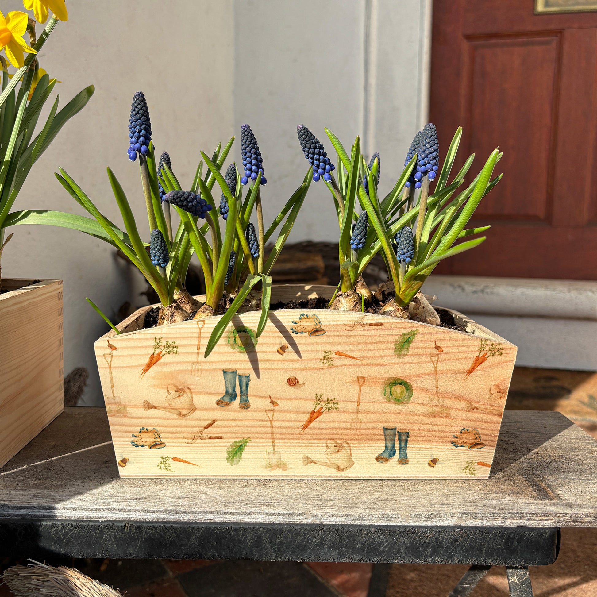 An arched wooden planter box full of spring flowers. The planter shows a watercolour repeat garden pattern design including welly boots, vegetables, gardening gloves, garden spade, snails, bees, robins and secateurs. A perfect gift for any occasion, especially for a garden enthusiast.