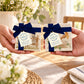 Two hands hold two sets of square marble coasters tied with dark blue ribbons and tags. The coasters have dark blue gift bows and ribbons with paper tags.