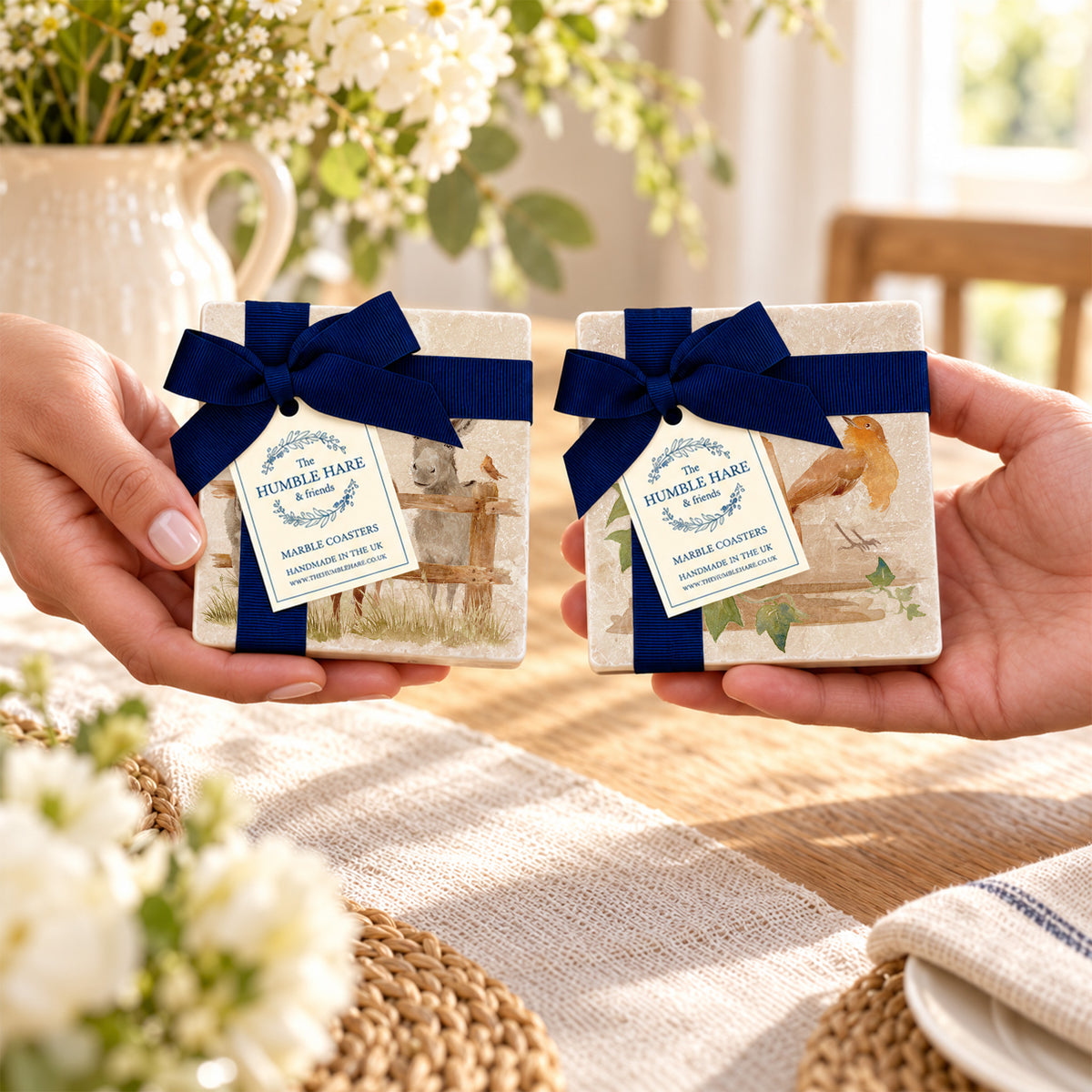 Two hands hold two sets of square marble coasters tied with dark blue ribbons and tags. The coasters have dark blue gift bows and ribbons with paper tags.