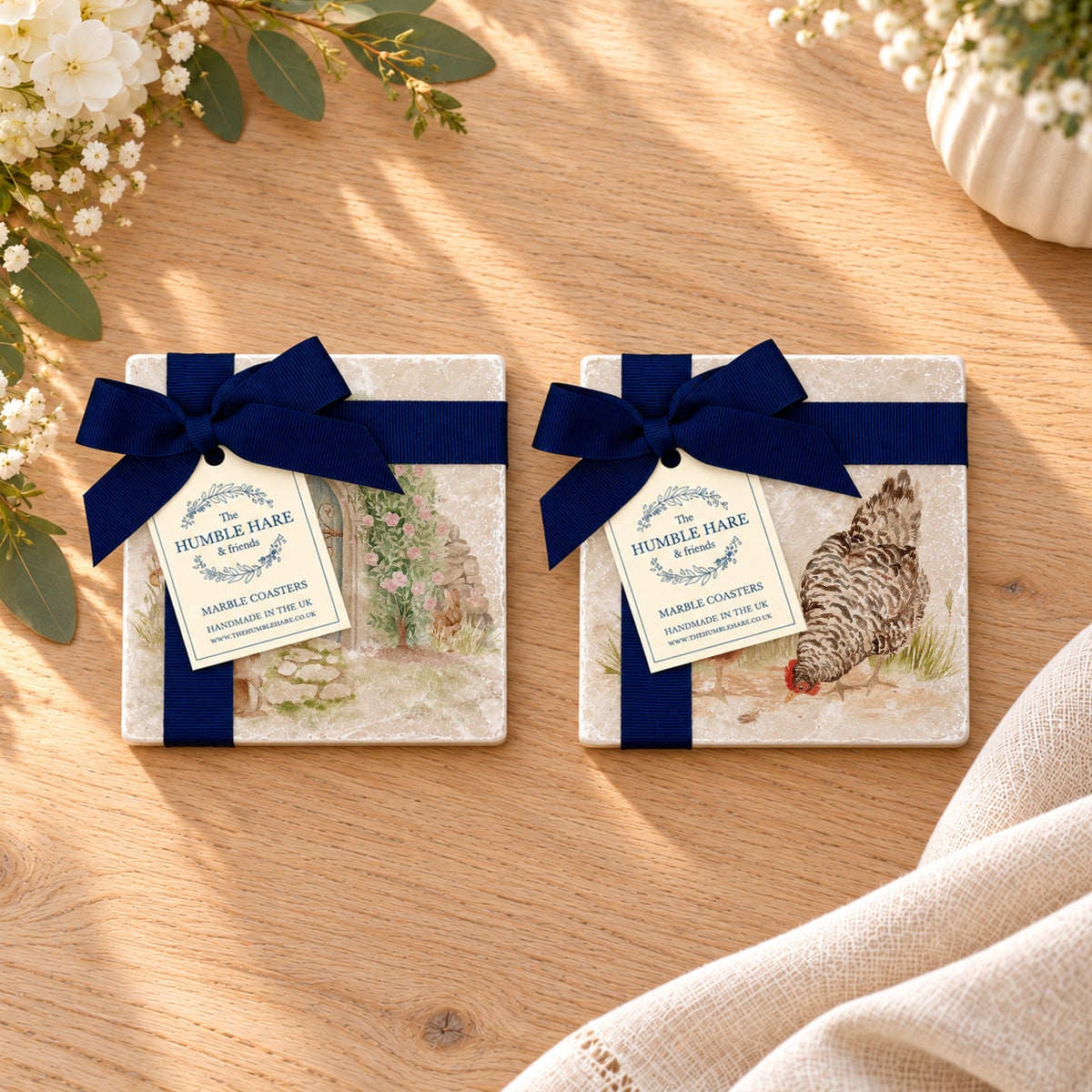 Two square marble coasters with decorative watercolour designs, tied with dark blue ribbons and hanging tags reading 'The Humble Hare,' arranged on a light wood surface.