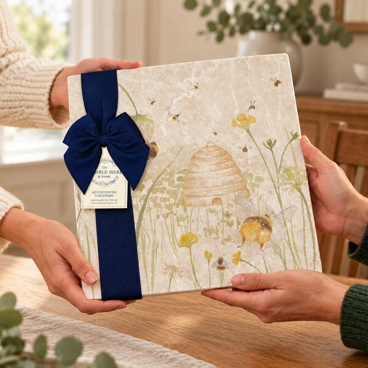 A large square marble trivet with a floral and bee illustration, wrapped in a wide navy ribbon with a large bow and a hanging tag, being held by two people at a wooden table in a warm indoor setting.
