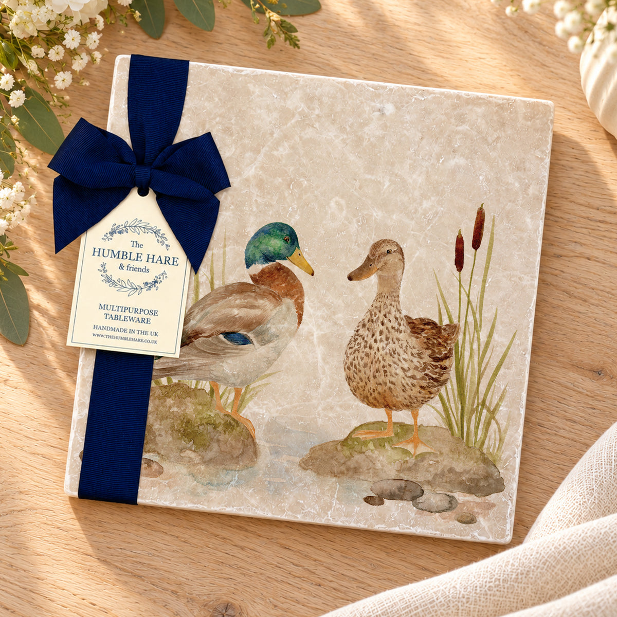 A large square marble placemat featuring a watercolour illustration of two ducks by a riverbank, tied with a dark blue ribbon and small paper tag. The item is photographed on a wooden surface with soft floral props around it.