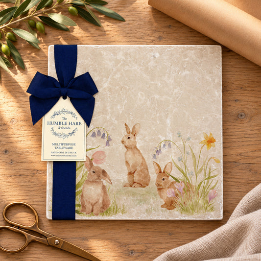 A square decorative marble platter with watercolour rabbits and flowers illustrations, and a dark blue ribbon with a paper tag reading 'The Humble Hare' It is styled on a wooden surface with scissors and a rolled sheet of brown wrapping paper.