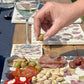 A sunny garden table set with marble platters and coasters that feature a watercolour grapevine pattern. The marble boards are being used to serve cheese and charcuterie.