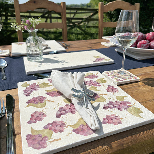 A large square marble placemat set on an outdoor table in the sunshine. The placemat features a watercolour pattern of grapes on the vine, and is decorated with a cream linen napkin.