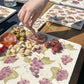 A set of marble serving platters and coasters on a sunny garden table, being used for charcuterie. The marble platters feature a watercolour pattern of grape vines.