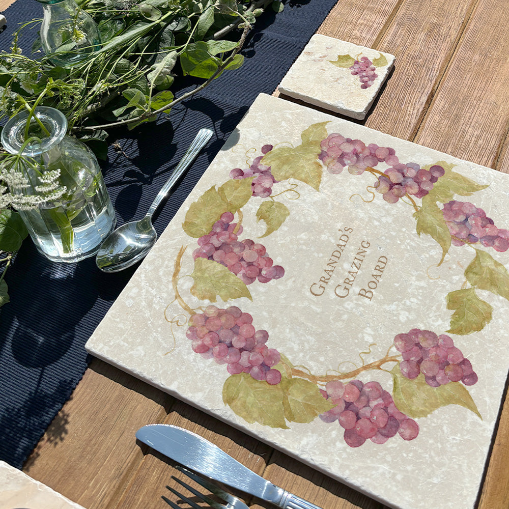 A personalised marble placemat with a grape vine and leaves wreath design, with a custom personalised message in the centre. The placemat is set on a garden table in the sunshine.