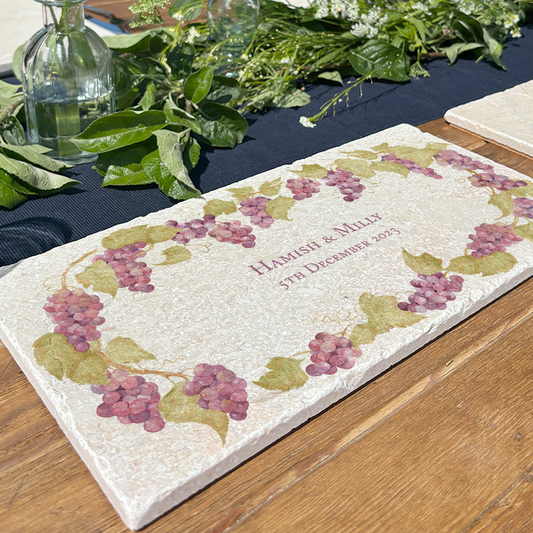 A personalised marble sharing board featuring a wreath of grape vines and leaves, with a bespoke message in the centre of the design with a couple’s names and anniversary date. The custom sharing platter is set on a sunny outdoor table in the garden.