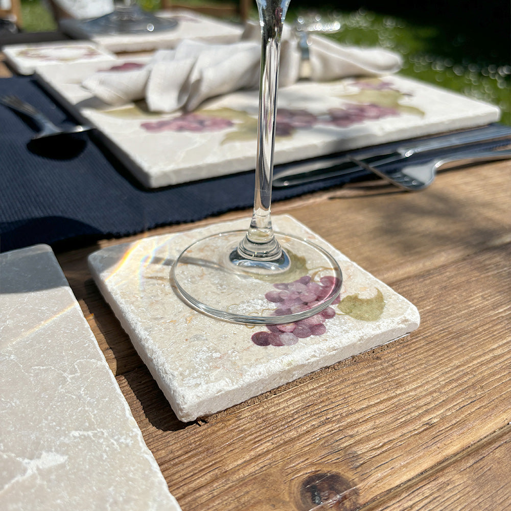 An al fresco dining table set with a wine glass on top of a square marble coaster featuring a minimalistic watercolour grape vine design.