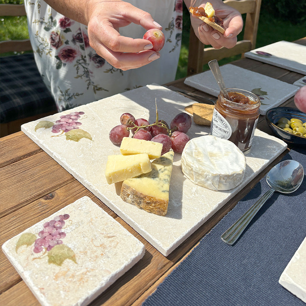 An al fresco dining table set with a marble cheeseboard and coasters featuring a minimalistic watercolour grape vine design.