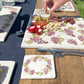 A square marble coaster featuring a watercolour grape vine wreath. The coaster is placed on a table set for an al fresco dinner with a charcuterie board and glasses of wine.