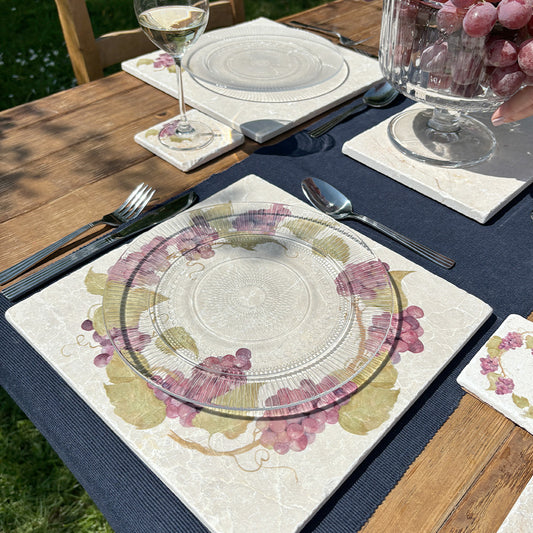 A garden table in the sunshine, the table is set with marble placemats and coasters featuring a rustic grape vine wreath design.