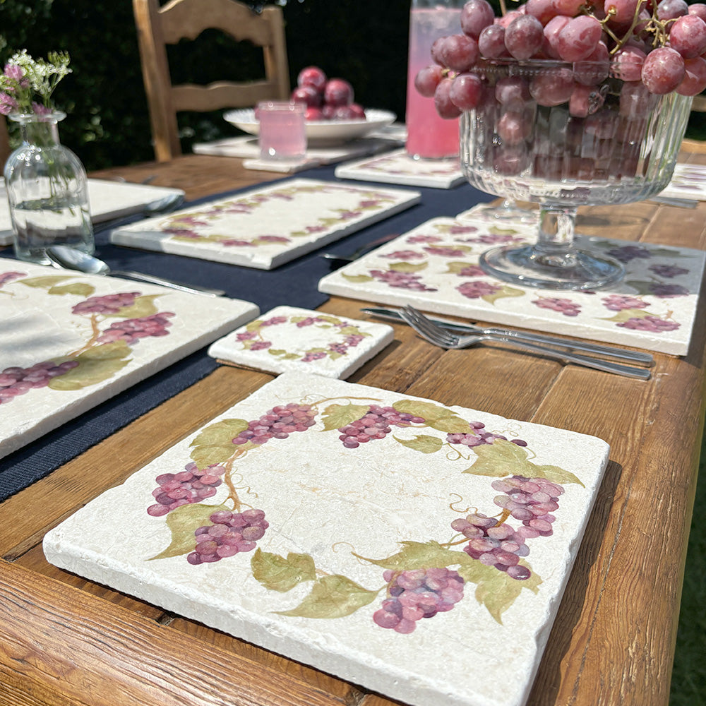 A medium square marble platter featuring a watercolour grape vine wreath design. The platter is set on a dinner table next to a marble placemat and coaster.