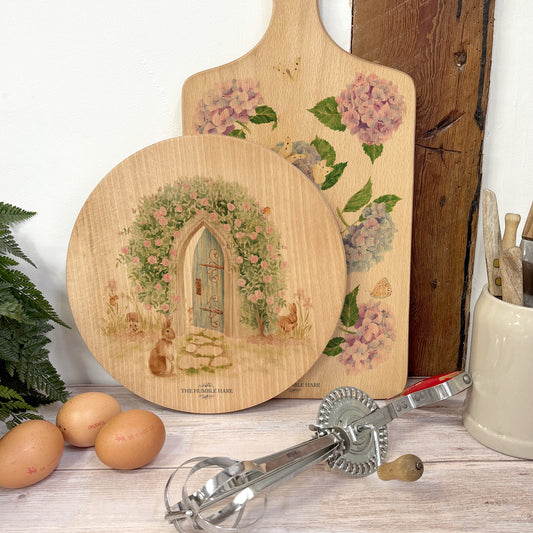 Round wooden chopping board on a kitchen worktop. The beech wood board features a watercolour design of a hidden garden with roses, rabbits and a hedgehog.