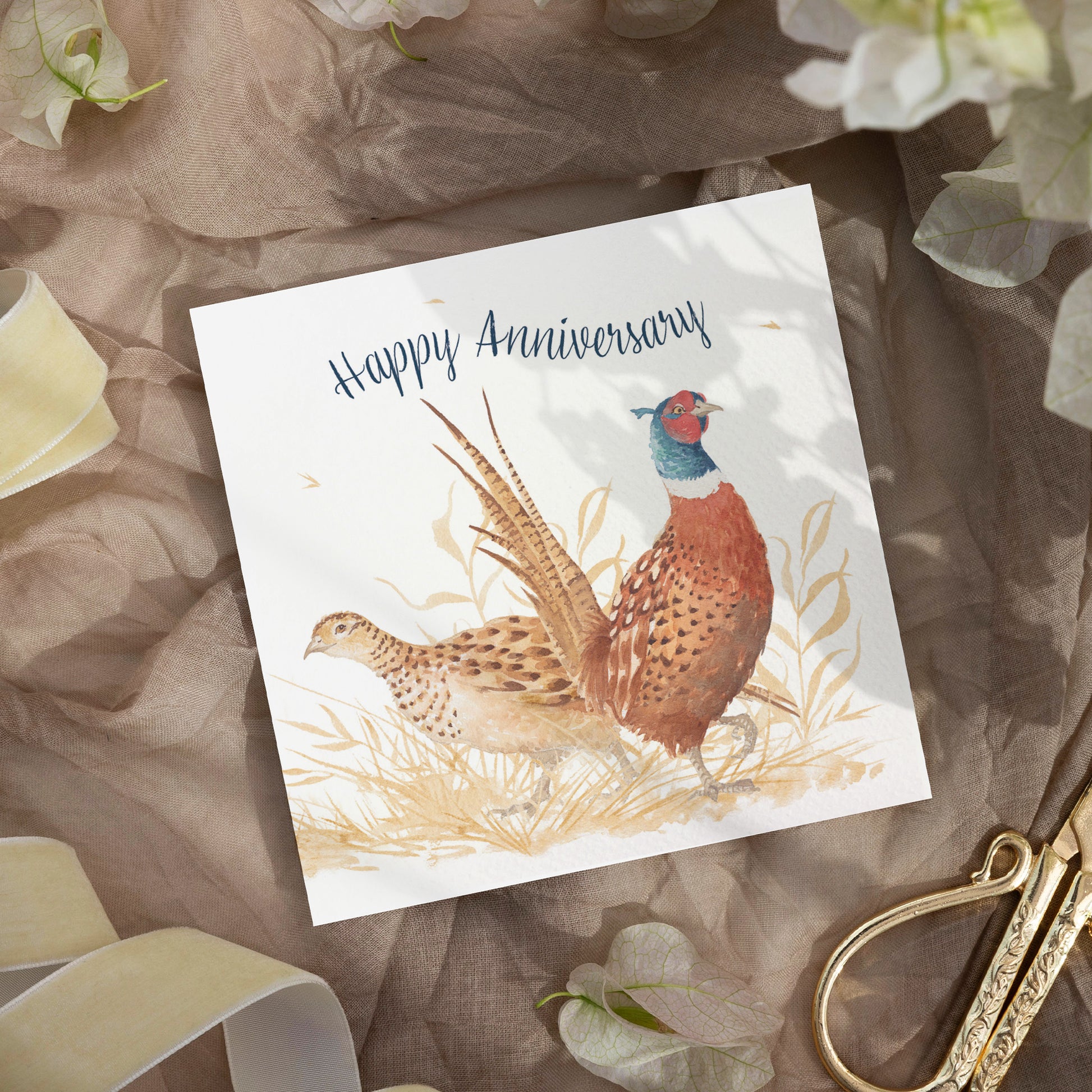 A greetings card laid flat on a table surrounded by gift wrapping items including scissors and ribbon. The card reads Happy Anniversary in dark blue text above a male and female pheasant couple in a watercolour style.