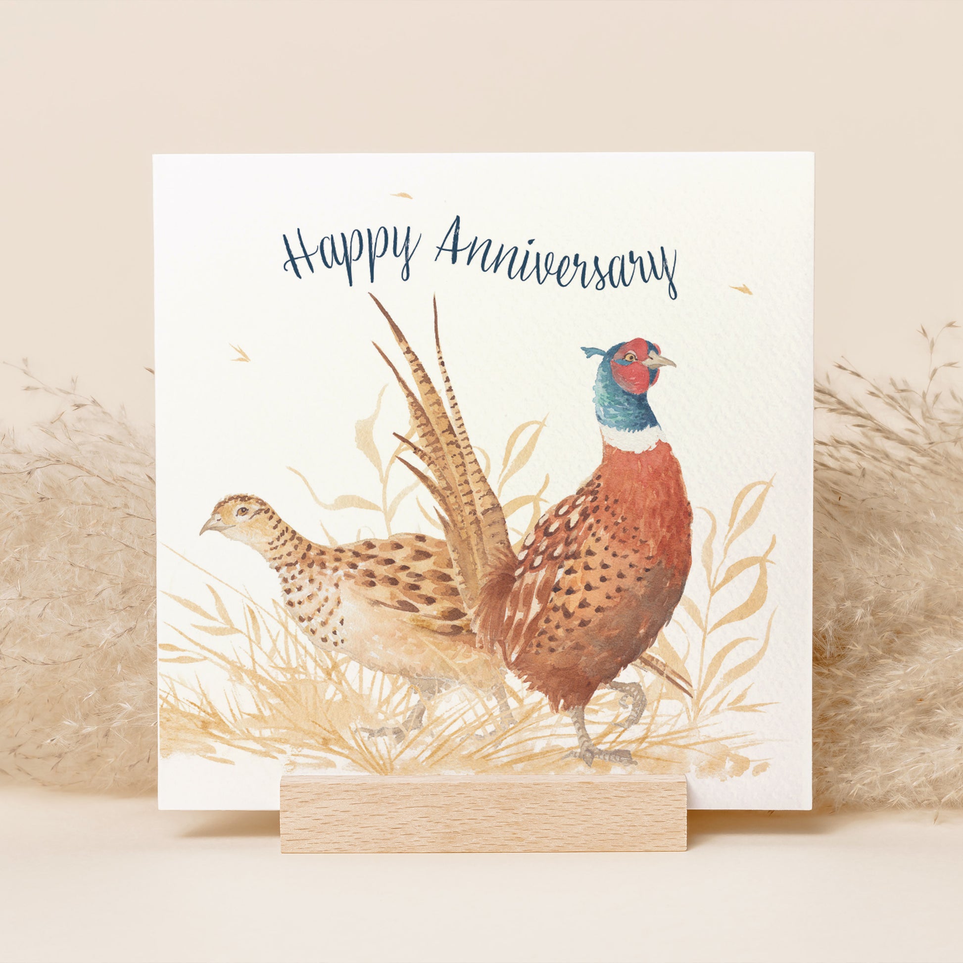 A greetings card reading Happy Anniversary in dark blue text above a male and female pheasant couple in a watercolour style.