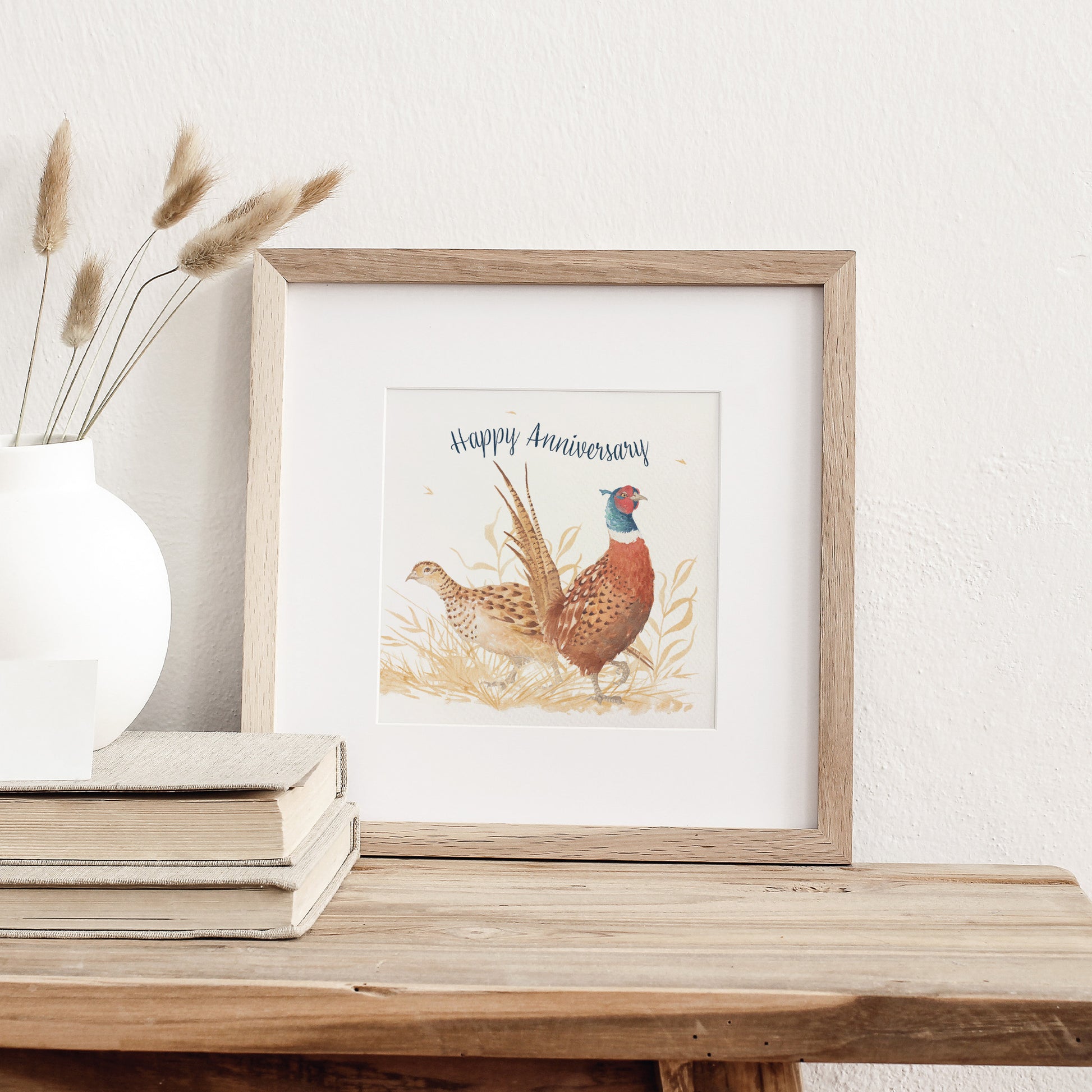 A greetings card displayed as an art print in a neutral coloured frame propped up on a bookshelf. The card reads Happy Anniversary in dark blue text above a male and female pheasant couple in a watercolour style.