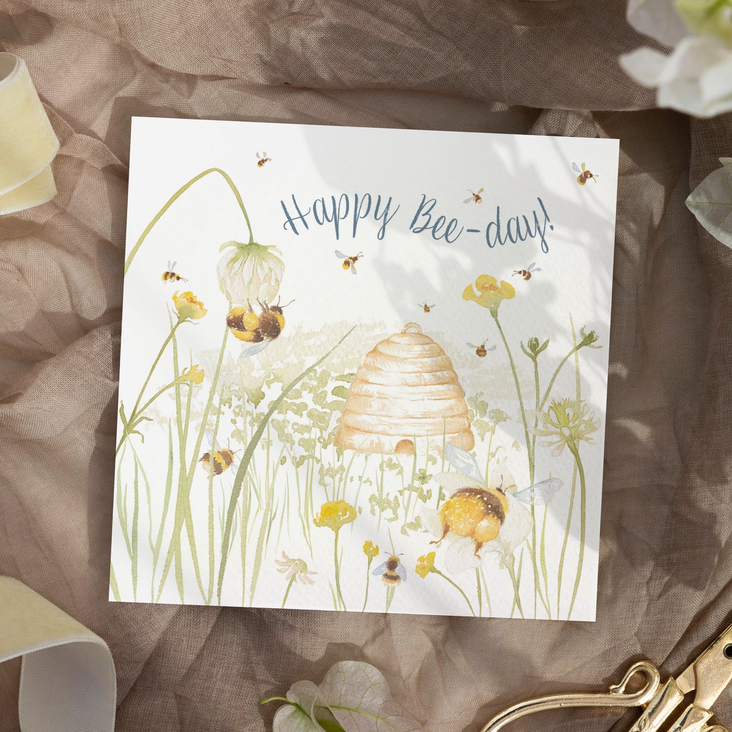 A greetings card laid flat on a table surrounded by gift wrapping items including scissors and ribbon. The card reads Happy Bee-day in dark blue text above a buttercup meadow full of bees around a traditional bee hive in a watercolour style.