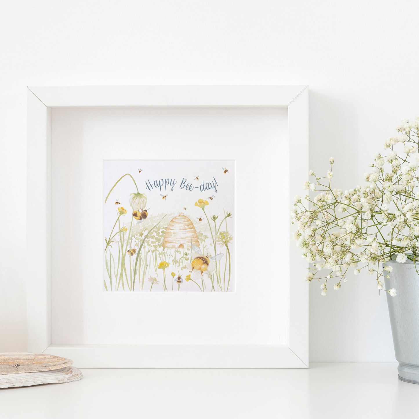 A greetings card displayed as an art print in a white frame propped up on a shelf next to a plant. The card reads Happy Bee-day in dark blue text above a buttercup meadow full of bees around a traditional bee hive in a watercolour style.