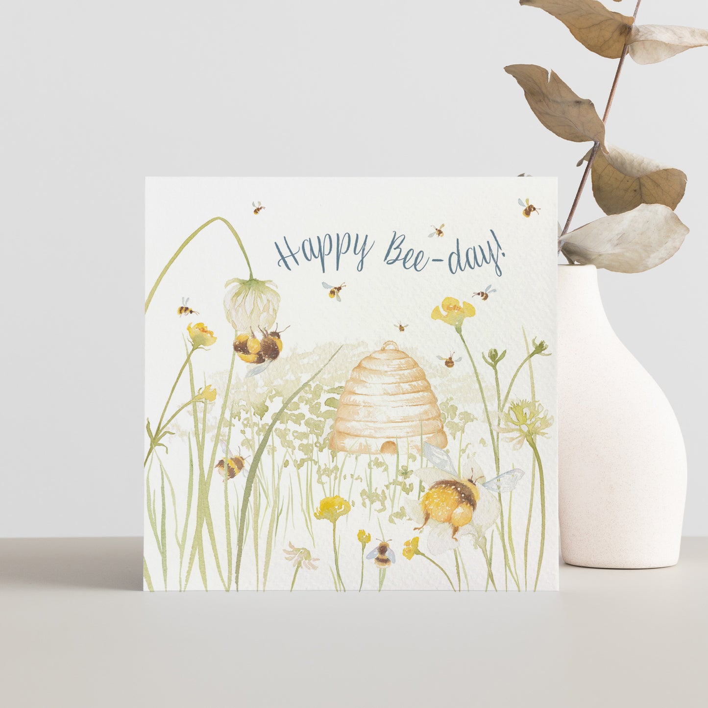 A greetings card reading Happy Bee-day in dark blue text above a buttercup meadow full of bees around a traditional bee hive in a watercolour style.