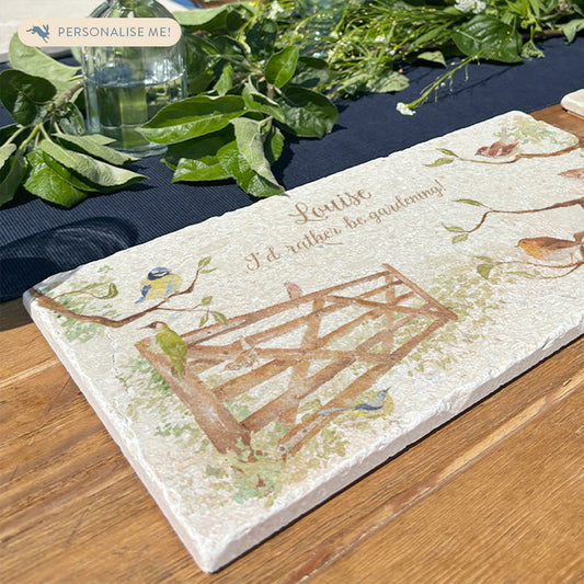 A personalised marble sharing platter set on a garden table, the platter features a gate surrounded by hedgerow with garden birds, and is personalised with a name and 'I'd rather be gardening!'.