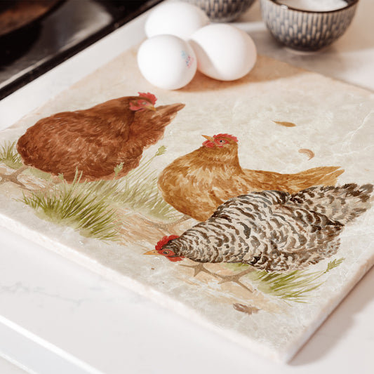 A large square marble platter featuring a watercolour farmyard scene of three hens. The platter is set on a kitchen worktop next to the hob to be used as a trivet.