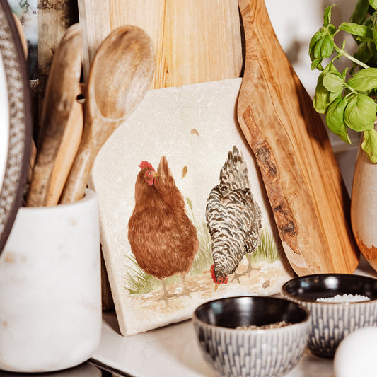 A medium marble platter propped up on a kitchen worksurface. The platter has a watercolour design of two hens.