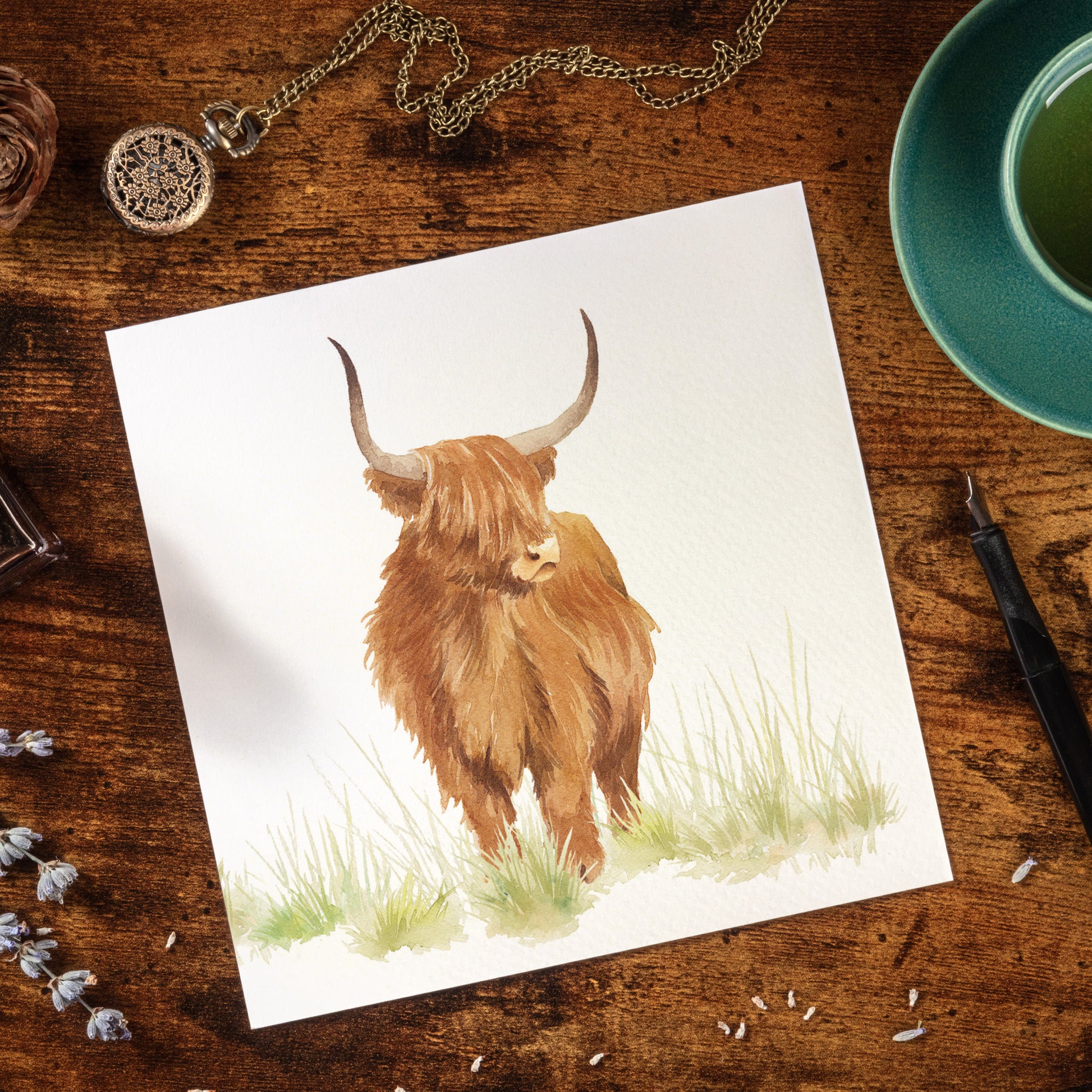 A greetings card laid flat on a table surrounded by writing items including a fountain pen and ink. The card features a highland cow in a watercolour style.