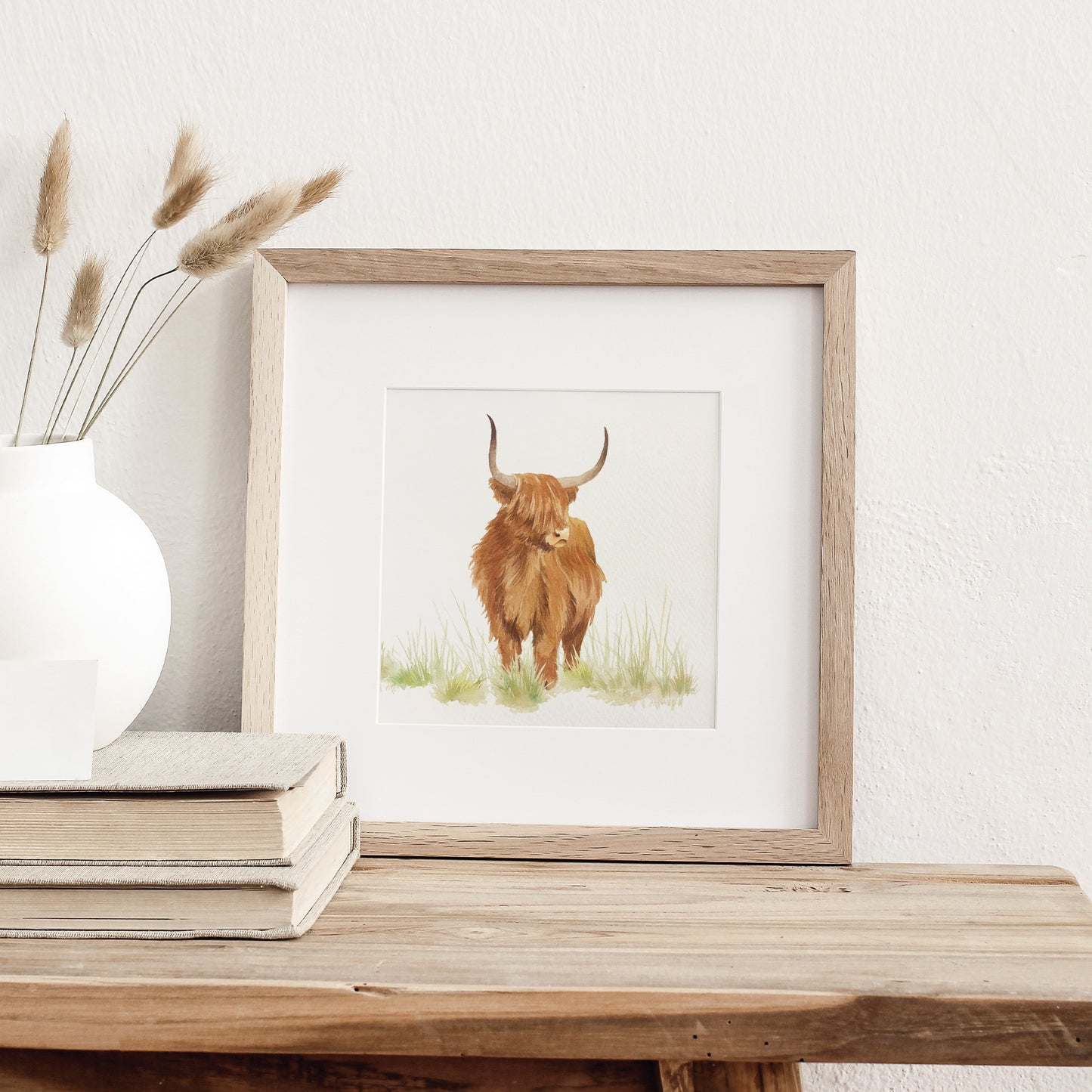 A greetings card displayed as an art print in a neutral coloured frame propped up on a bookshelf. The card features a highland cow in a watercolour style.