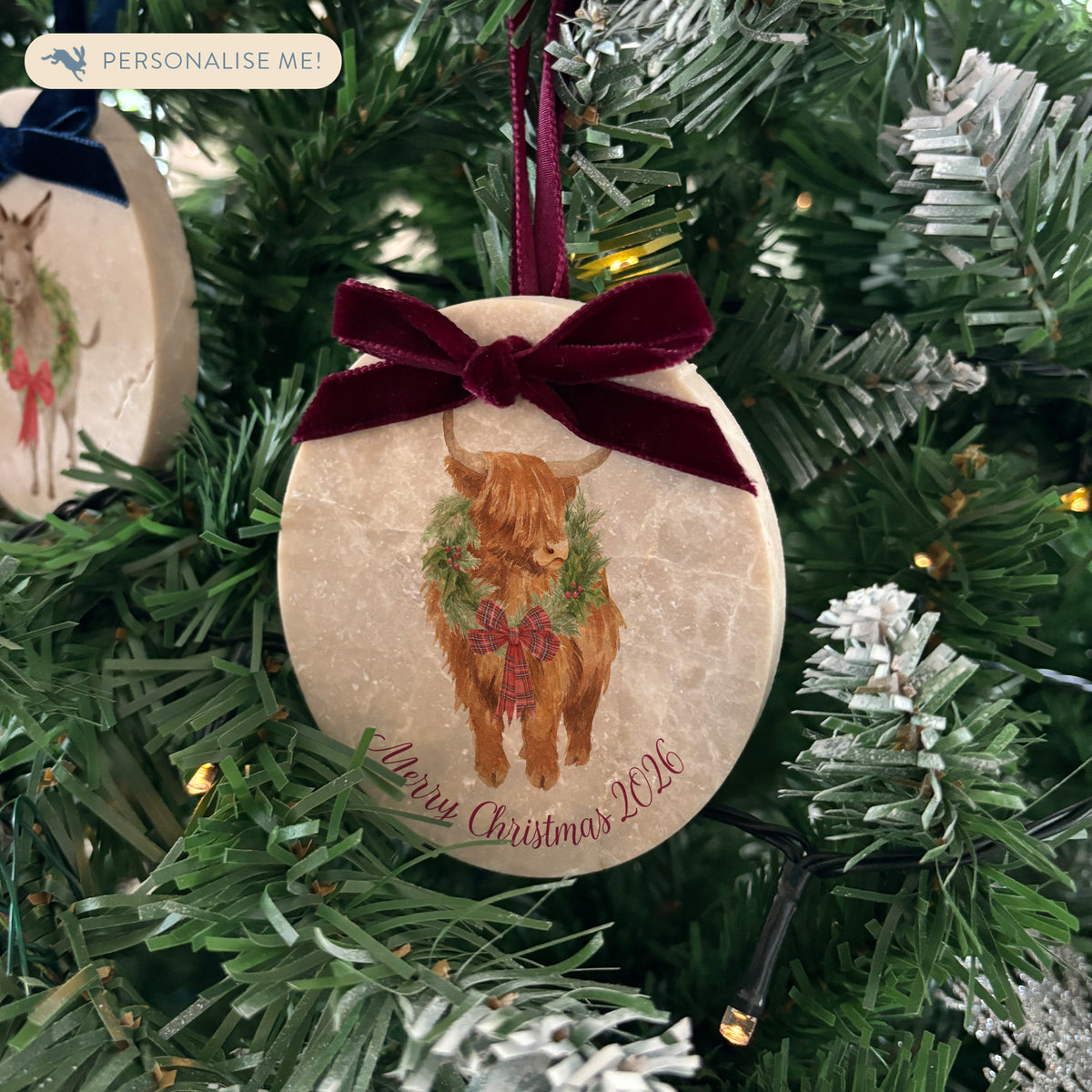 An oval marble Christmas ornament with a red velvet ribbon loop and bow hanging on a Christmas tree, featuring a vintage-style illustration of a highland cow wearing a green wreath and red bow, with personalised text written at the bottom.