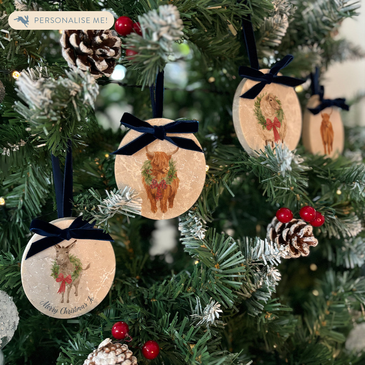 Four oval marble Christmas ornaments with blue velvet ribbon loop and bows hanging on a Christmas tree, featuring vintage-style illustrations of a robin, highland cow, sheep and donkey wearing a green wreath and red bow, with personalised text written at the bottom.
