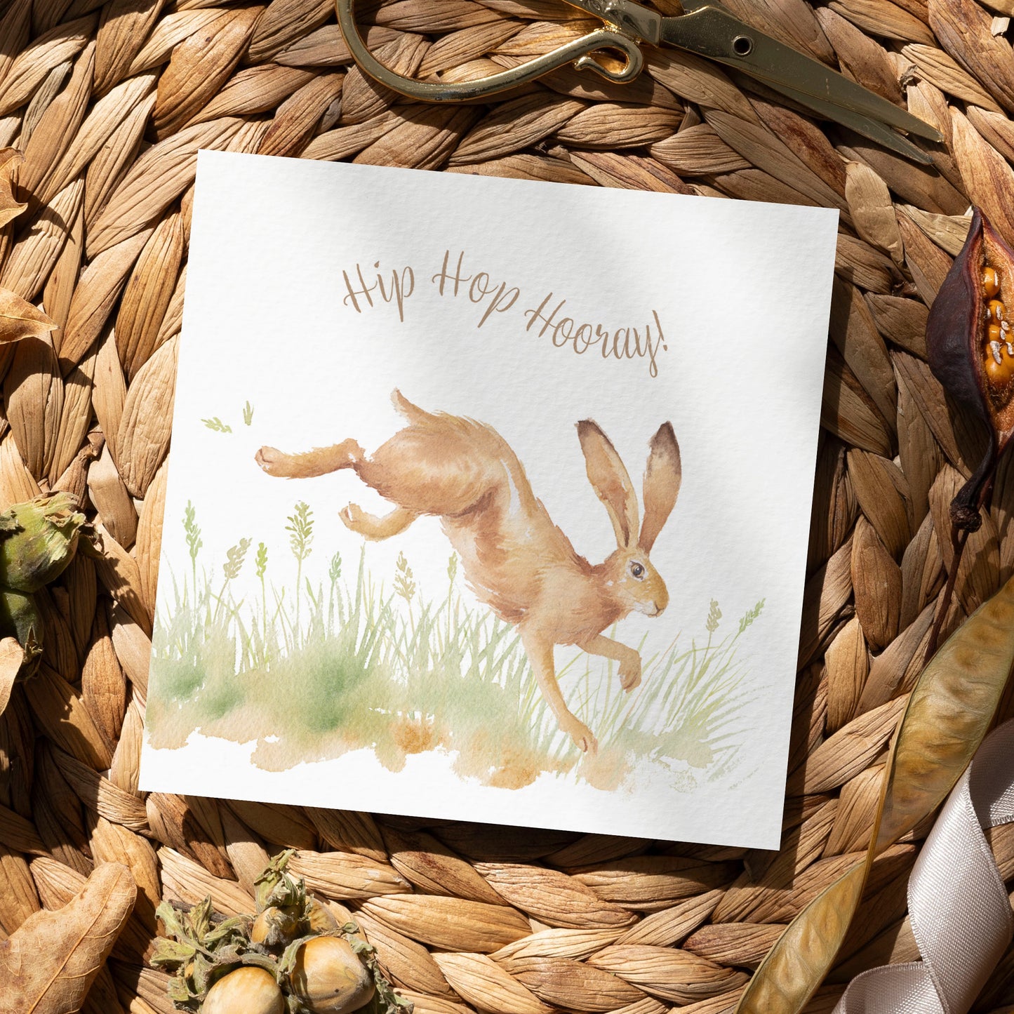 A greetings card laid flat on a table surrounded by gift wrapping items including scissors and ribbon. The card reads Hip Hop Hooray in brown text above a leaping hare in a watercolour style.
