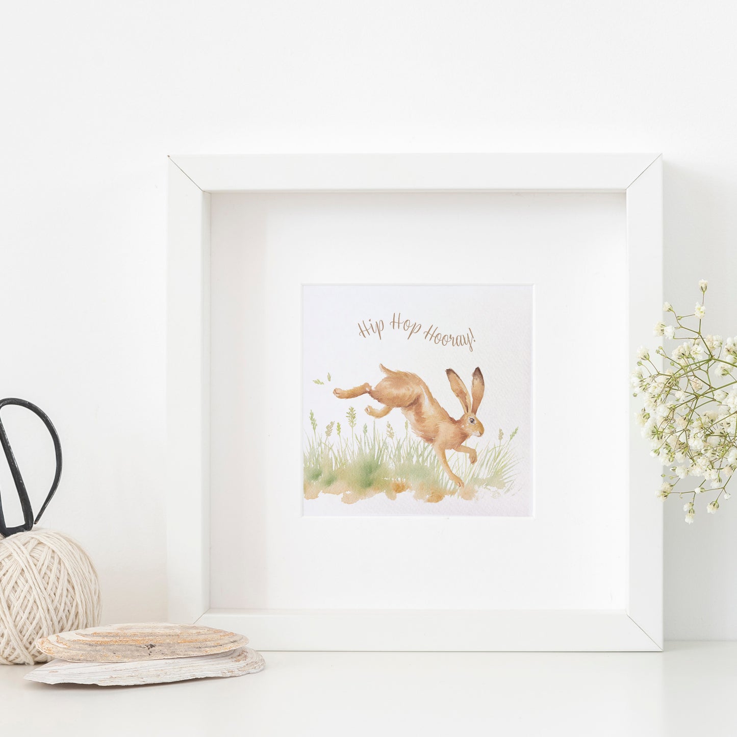 A greetings card displayed as an art print in a white frame propped up on a shelf next to a plant. The card reads Hip Hop Hooray in brown text above a leaping hare in a watercolour style.