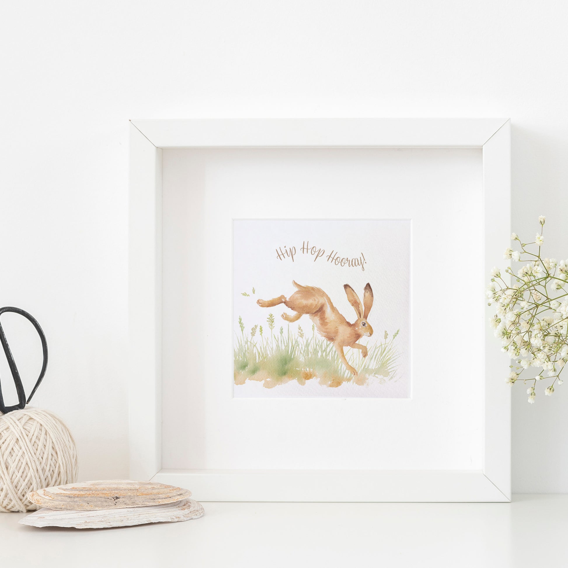 A greetings card displayed as an art print in a white frame propped up on a shelf next to a plant. The card reads Hip Hop Hooray in brown text above a leaping hare in a watercolour style.