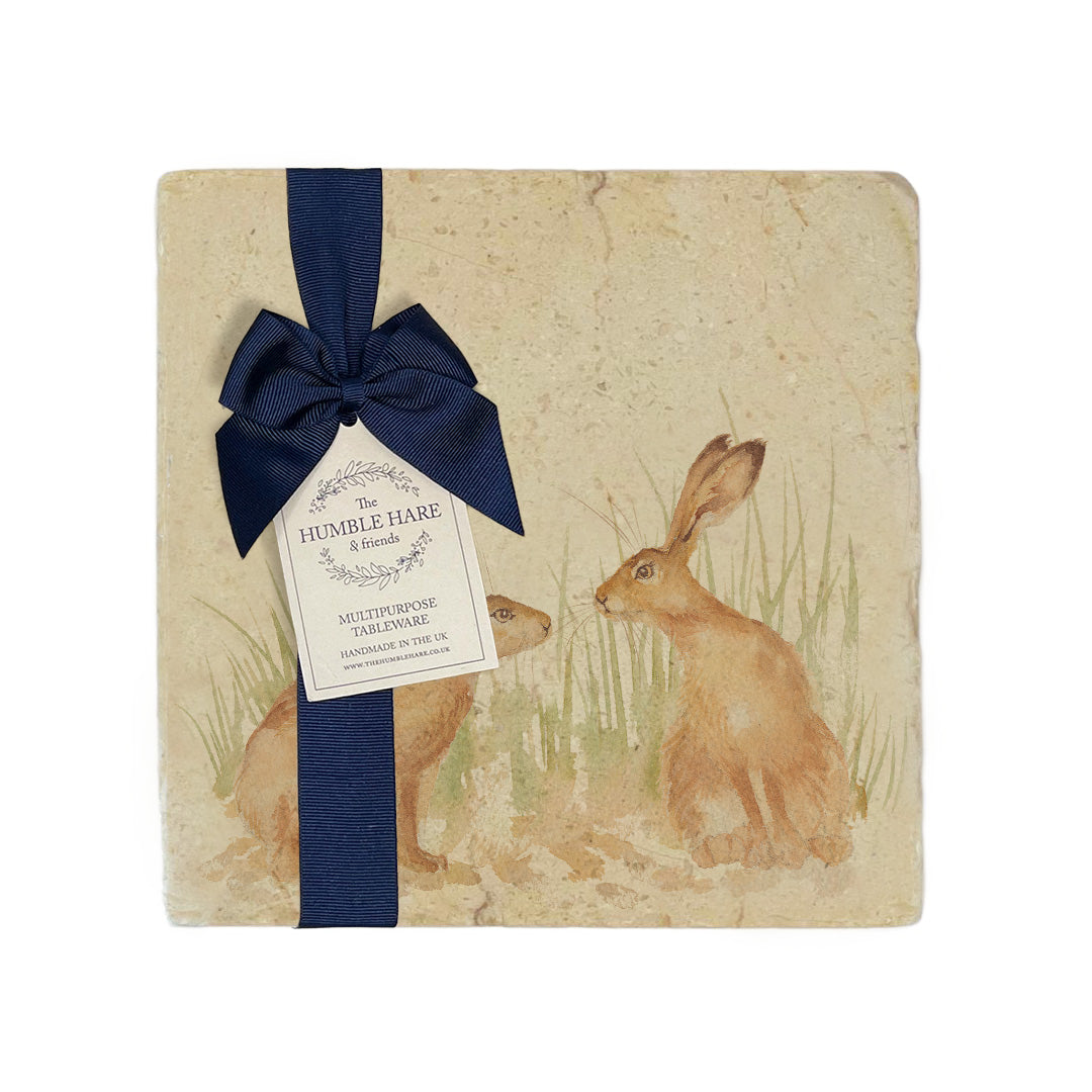 A multipurpose marble platter with a design featuring two hares facing each other about to touch noses, packaged with a luxurious dark blue bow and branded gift tag.