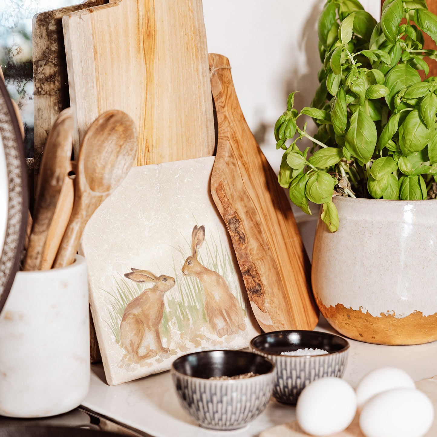 A medium square cream marble platter propped up on a country kitchen worksurface among kitchen utensils and wooden chopping boards. The marble platter features a watercolour design of two hares facing each other about to touch noses.