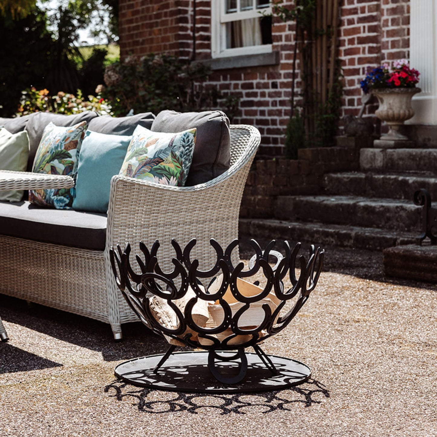A handmade horseshoe fire pit next to a seating area in the garden of a country house. The fire pit has been filled with firewood and has a black painted finish.