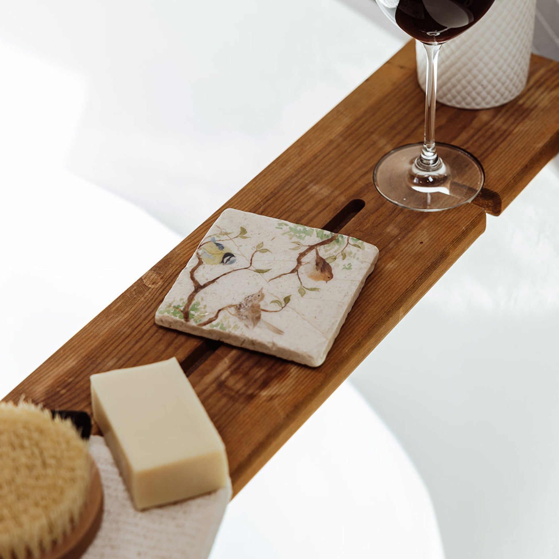 A square marble coaster on a wooden bath tidy above a bubble bath. The coaster features a watercolour design of garden birds in the hedgerow around a paddock gate.