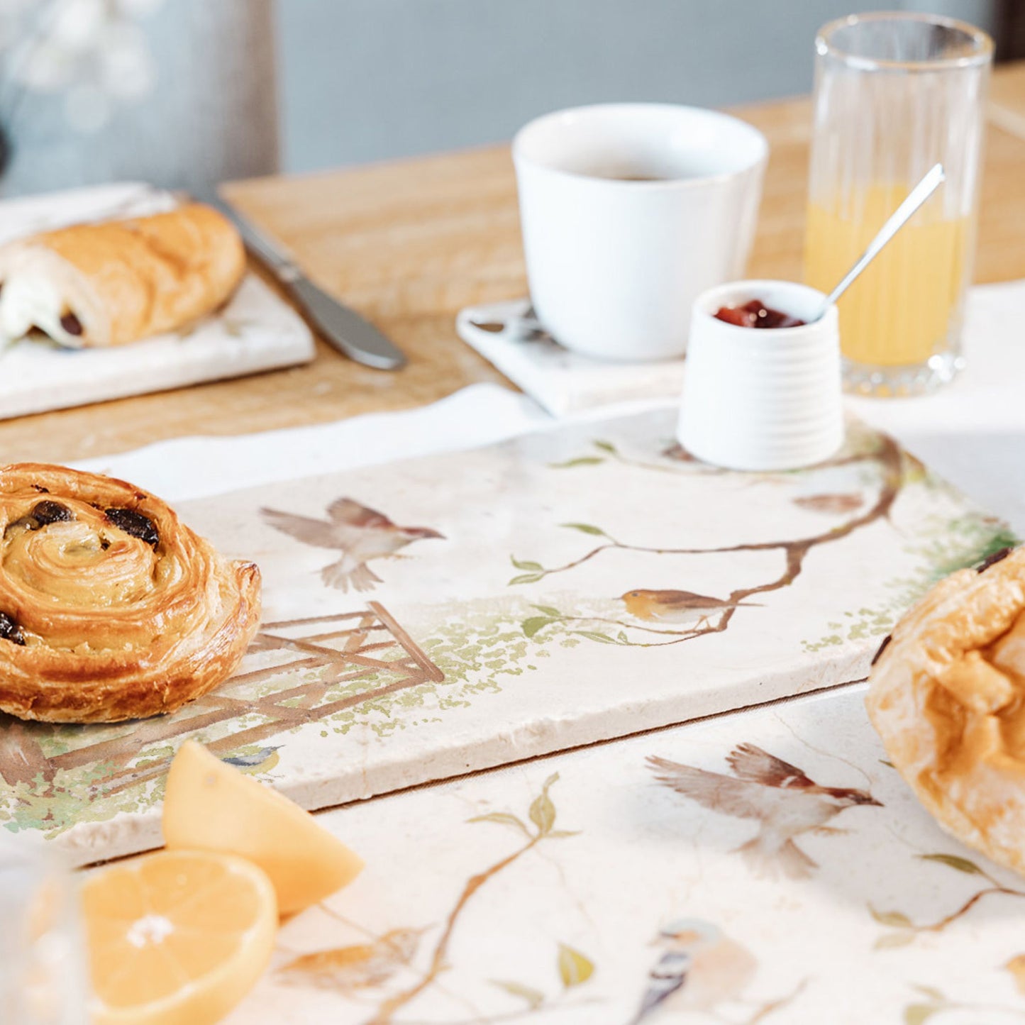 A breakfast table set with a marble sharing platter featuring British garden birds in the hedgerow around the paddock gate, in a watercolour style. The platter is being used to serve pastries and fruit.