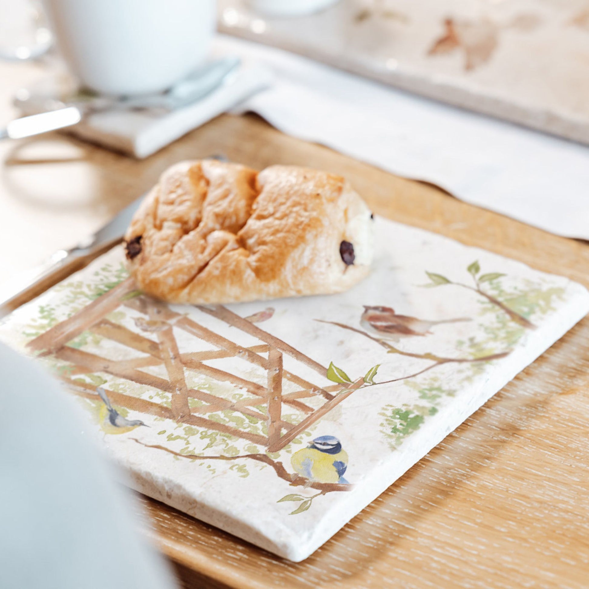 A medium multipurpose marble platter with a watercolour design featuring British garden birds in the hedgerow around the paddock gate. The platter is being used as a plate for breakfast pastries.