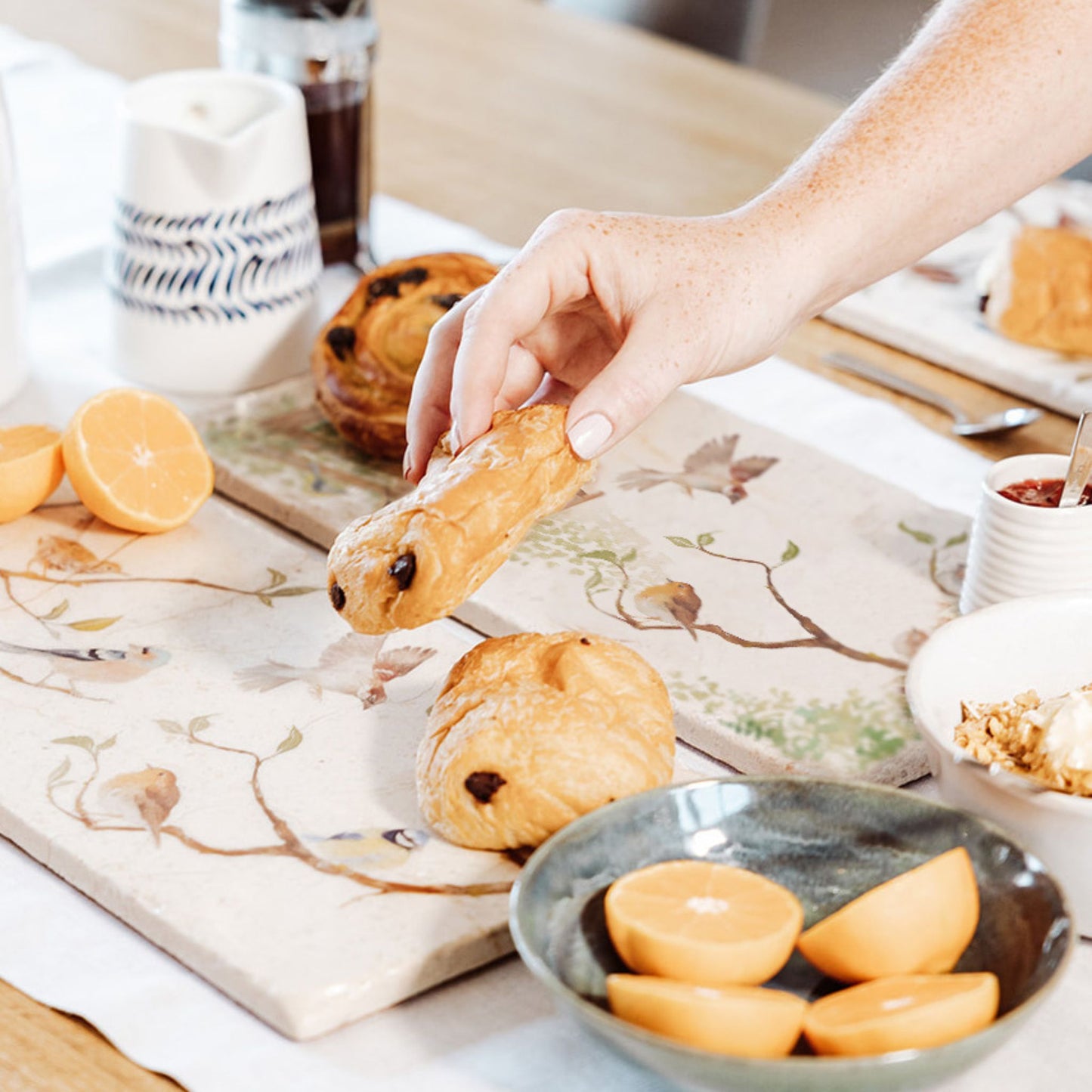 A breakfast table set with a marble sharing platter featuring British garden birds in the hedgerow around the paddock gate, in a watercolour style. The platter is being used to serve pastries and fruit.