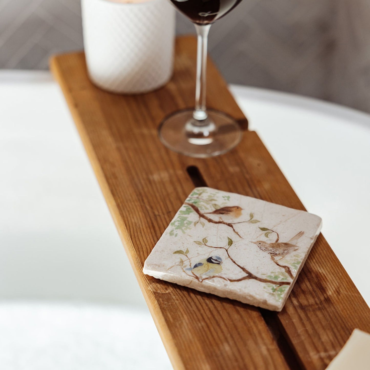 A square marble coaster on a wooden bath tidy above a bubble bath. The coaster features a watercolour design of garden birds in the hedgerow around a paddock gate.