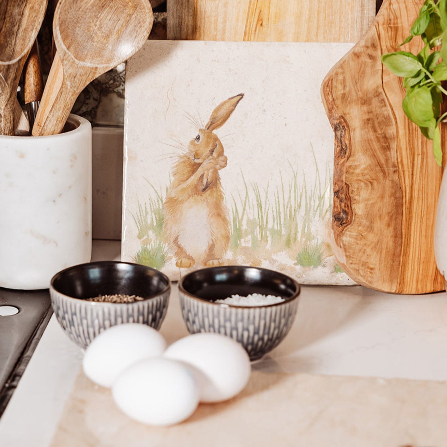 A medium square cream marble platter propped up on a country kitchen worksurface among kitchen utensils and wooden chopping boards. The marble platter features a watercolour design of a hare washing his ear.