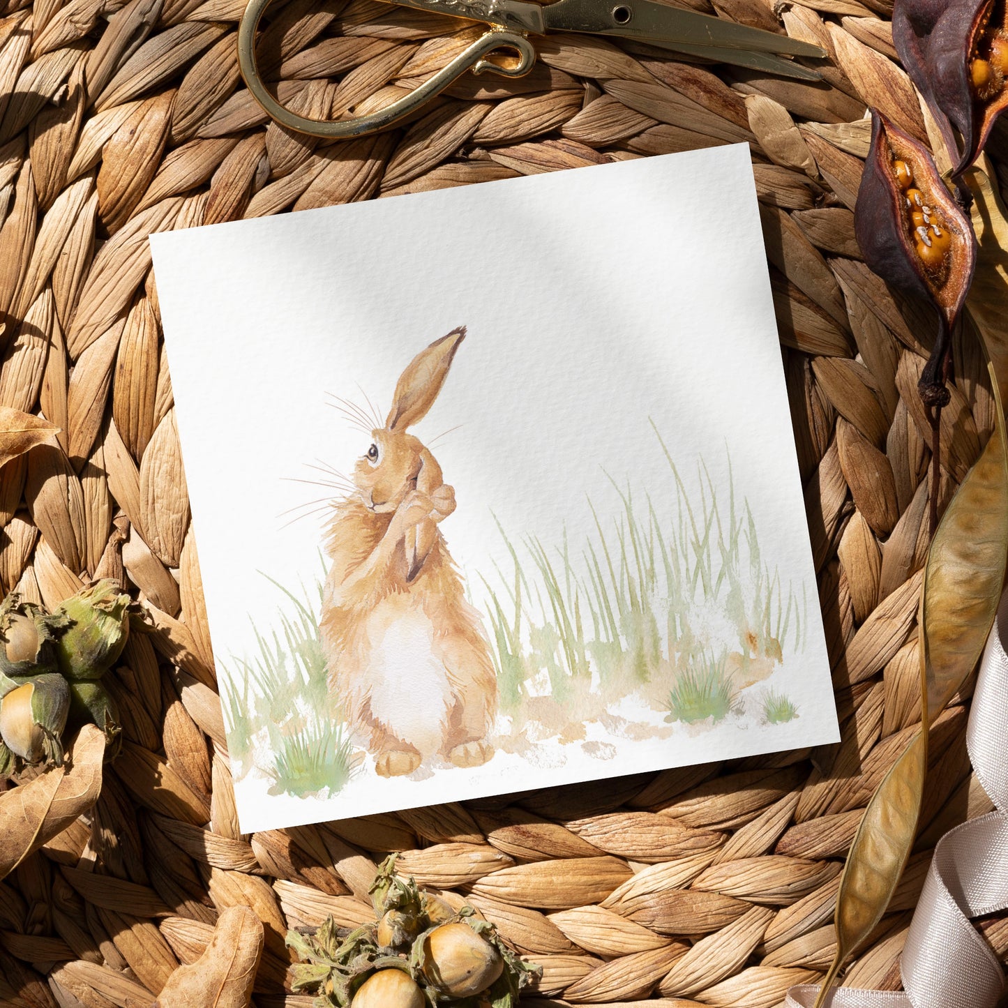A greetings card laid flat on a table surrounded by gift wrapping items including scissors and ribbon. The card features a hare washing his ear in a watercolour style.