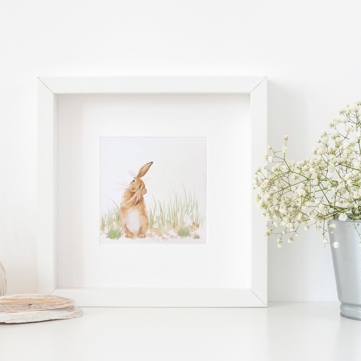 A greetings card displayed as an art print in a white frame propped up on a shelf next to a plant. The card features a hare washing his ear in a watercolour style.